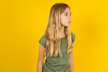 blonde kid girl wearing green T-shirt over yellow studio background stares aside with wondered...