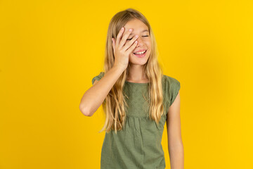 blonde kid girl wearing green T-shirt over yellow studio background makes face palm and smiles...