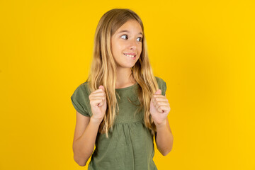 blonde kid girl wearing green T-shirt over yellow studio background clenches fists and awaits for...