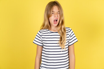 young beautiful blonde kid girl over yellow studio background yawns with opened mouth stands. Daily...