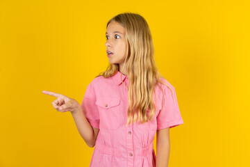 Stunned young beautiful blonde kid girl over yellow studio background with greatly surprised expression points away on copy space, indicates something