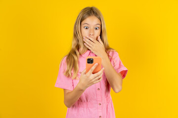 young beautiful blonde kid girl over yellow studio background being deeply surprised, stares at smartphone display, reads shocking news on website, Omg, its horrible!