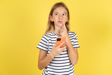 young beautiful blonde kid girl over yellow studio background thinks deeply about something, uses...