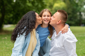 Portrait of a Lovely Mixed Race Family where Mom and Dad are Kissing their Blond Daughter on the Cheek. Beautiful day for mutual hugs and congratulations. Family rests at the weekend in the park.