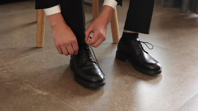 A man in a suit ties up shoelaces on black leather shoes on a wooden parquet background business style. preparation for a wedding or business meeting. means for care of leather shoes
