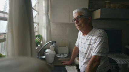 Thoughtful Senior Man Gazing Out of Kitchen Window, Leaning on Counter. Retired older person thinking while staring out window - obrazy, fototapety, plakaty
