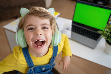 Smiling Primary school Student child with headphones posing using laptop with green screen chroma...