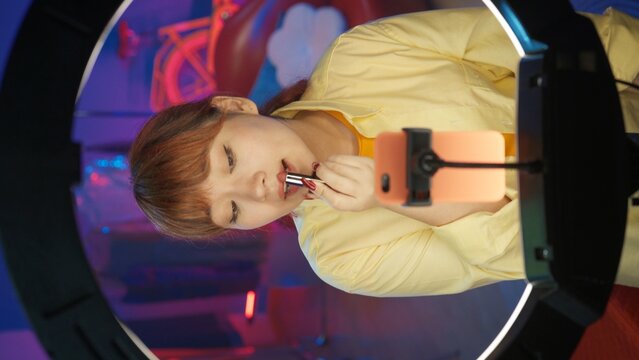 Attractive girl vlogger in casual clothing applying red lipstick in front of the camera at home studio. Vertical photo