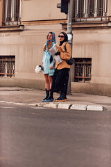 Couple woman one wearing a hijab and a modern yet traditional dress, and the other in a blue dress and scarf, walking together through the city at sunset. One carries a bouquet and bread, while the