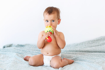 a small baby of 10 months in a diaper sits on a bed with a rattle and looks at the camera