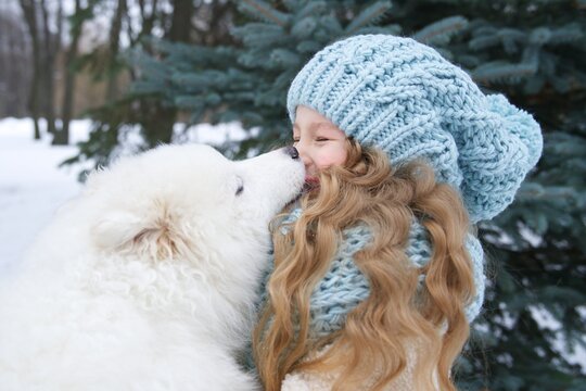 Little beautiful girl, cute happy positive child kid is lying on snow, play, having fun with her big white dog at winter day in park or forest