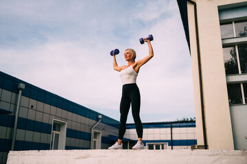 Caucasian blonde woman doing weight workout with dumbbells outdoors. Healthy lifestyle, muscular build woman. 