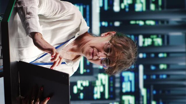 Vertical video Senior computer scientist in server farm completing tasks on tablet while running scripts on laptop terminal. Expert amidst supercomputers manipulating code and improving cybersecurity