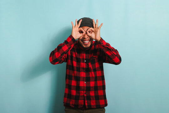 Excited young Asian man with beanie hat and red plaid flannel shirt makes the OK sign gesture with his fingers, acting as binoculars with his eyes peeking through them, isolated on blue background - Powered by Adobe