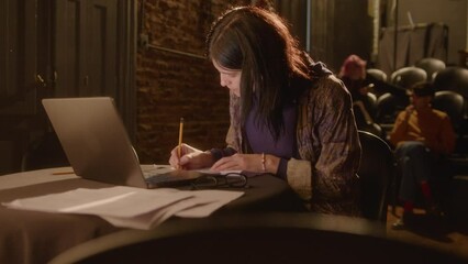 Professional female playwright working with laptop, writing down notes, smiling and posing for camera on stage in a theater. Medium long shot, video portrait