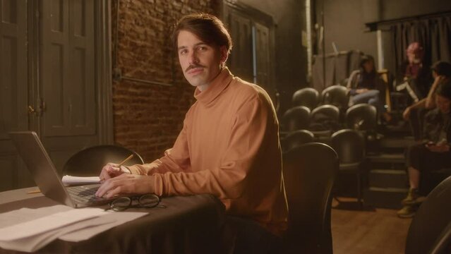 Professional male playwright sitting at table with laptop and papers and posing for the camera with smile, working on stage in a theater. Medium long shot, video portrait