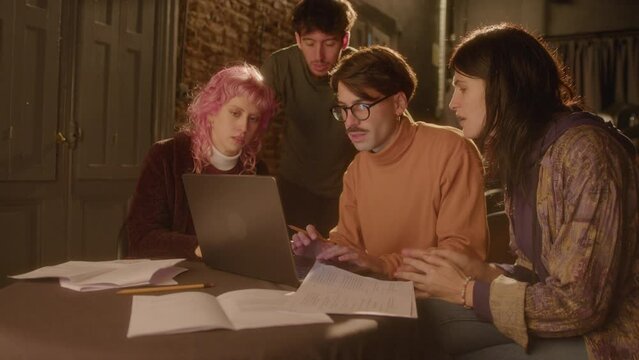Stage director and group of young actors discussing something on laptop and working on play script together at table with lots of papers while preparing for theater performance