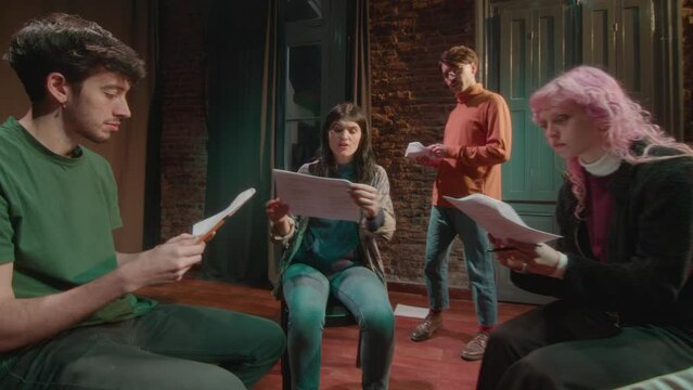 Young drama students sitting on theater stage and reading casting monologues from papers, director walking around and giving advices
