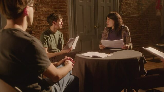 Company of drama students reading printed play scripts and discussing their monologues with stage director while preparing for performance in a theater