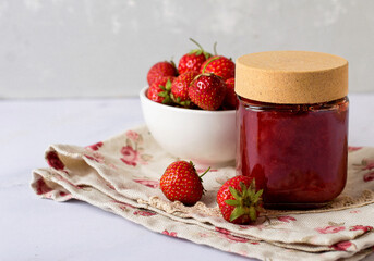 A jar of strawberry jam, a bowl of strawberries on the kitchen table.