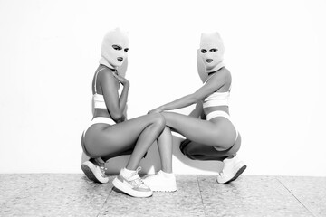Two beautiful sexy women in underwear. Models wearing bandit balaclava mask. Hot seductive female in nice lingerie posing near white wall in studio. Crime and violence. Sits on floor