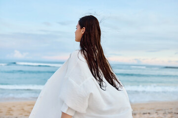 Happy tanned slender woman in white swimsuit shirt and denim shorts walks on the beach with her back to the camera on the sand by the ocean with wet hair after swimming, sunset light