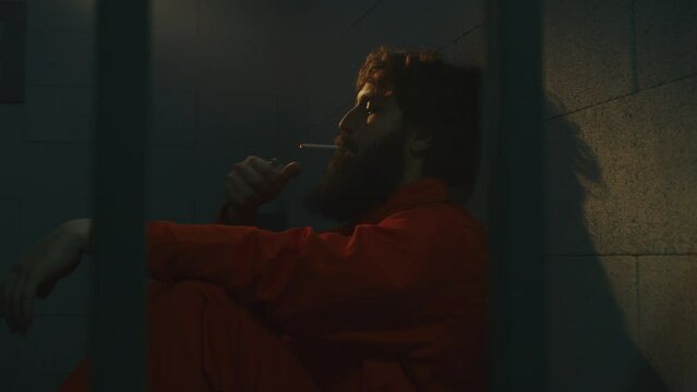 Depressed male prisoner in orange uniform sits on the bed, smokes cigarette in prison cell. Criminal, inmate serves imprisonment term for crime in jail. Detention center or correctional facility.
