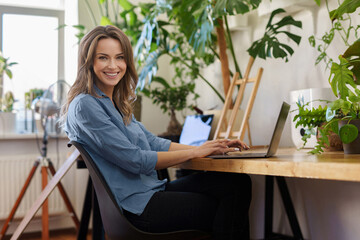 A charming smiling freelance woman in a blue shirt working on her laptop in a plant-filled room