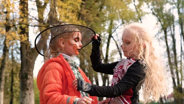 A girl dressed as a cheerleader and with half-face makeup in the form of a skeleton adjusts her hat to a girl in the form of a pumpkin. Horizontal video