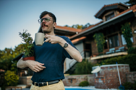 Shot of a man standing in front of his house, holding a plant and drinking a coffee.