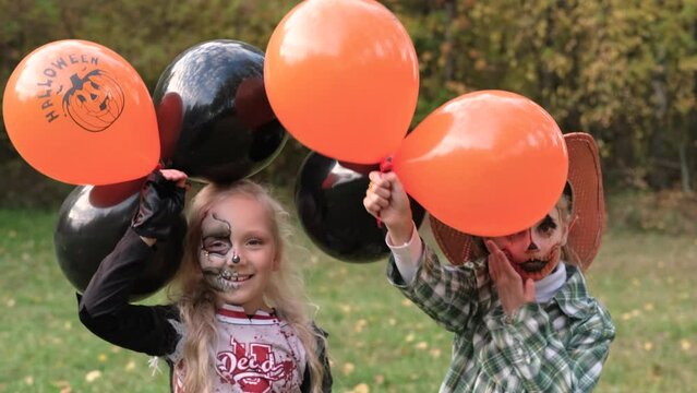 Two children girls girlfriends in Halloween costumes and make-up make frightening grimaces against the background of autumn leaves and with balloons in their hands. Horizontal video