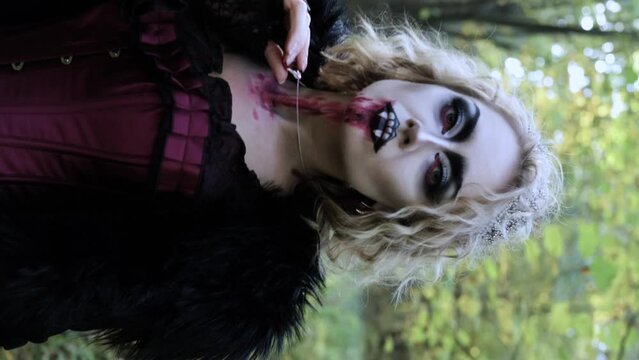 A woman in the image of a vampire or a witch with white make-up and in a medieval dress poses in the forest in front of the camera. Vertical video