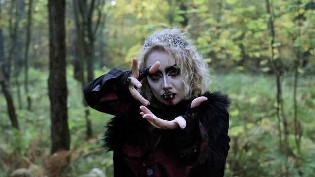 A woman in the image of a vampire or a witch with white make-up and in a medieval dress poses in the forest in front of the camera. Horizontal video