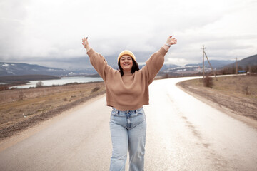 Beutiful woman is wearing jeans, sweater and hat walking in cold weather along the road        against the background of landscape and nature. A plus size girl is travelling - obrazy, fototapety, plakaty