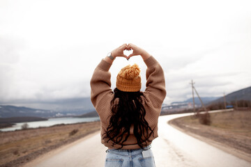 back view overweight model makes heart. Beutiful woman is wearing jeans, sweater and hat walking in...
