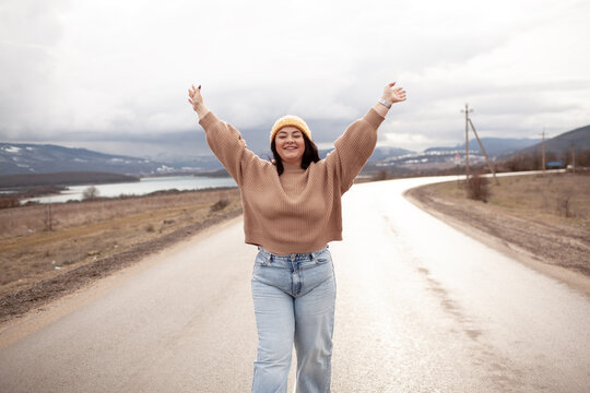 Beutiful woman is wearing jeans, sweater and hat walking in cold weather along the road        against the background of landscape and nature. A plus size girl is travelling