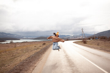 Beutiful woman is wearing jeans, sweater and hat walking in cold weather along the road        against the background of landscape and nature. A plus size girl is jumping