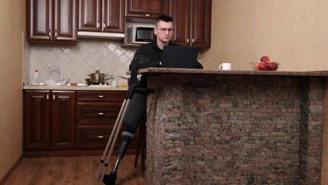 disabled worker, artificial limb, independent living. Man wearing glasses, prosthetic leg, operates laptop while crutches are nearby.