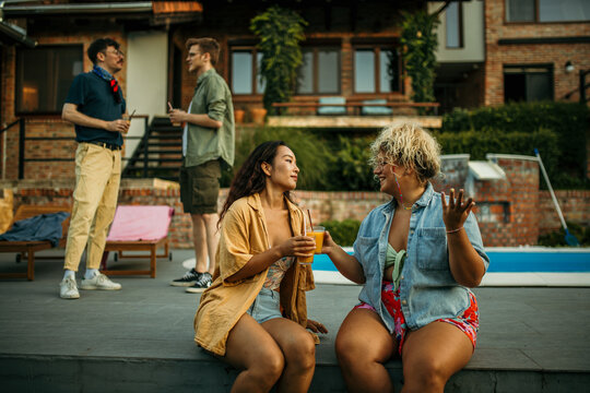 Two diverse female friends sitting and drinking cocktails, men standing and talking in the background during a house pool party.