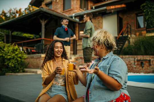 Two diverse female friends sitting and drinking cocktails, men standing and talking in the background during a house pool party.