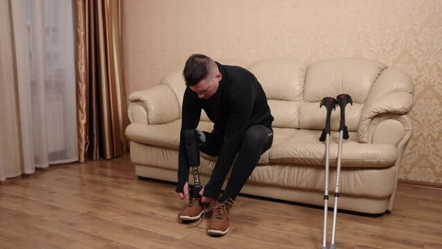 disabled man, prosthetic technology, leg amputation. With his crutches lying nearby, man who uses prosthetic leg due to disability is sitting on couch and putting on shoes.