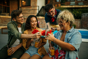 Gropu of people sitting and eating watermelon slices by the pool.