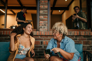 Two females sit and talk, while men prepare food and wine for them at home.
