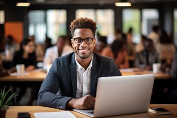 Fototapeta premium Confident young African businessman sitting at a desk with a group of colleagues in the background working on the laptop computer