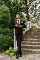 A man in a classic black suit and white shirt stands and poses near the stone steps. A stylish...