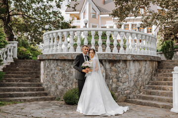 Portrait of a young bride in a white dress and a brunette groom in a suit on the stairs embracing near an old city park. A beautiful and romantic wedding, a happy couple