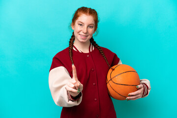 Young reddish woman playing basketball isolated on blue background showing and lifting a finger