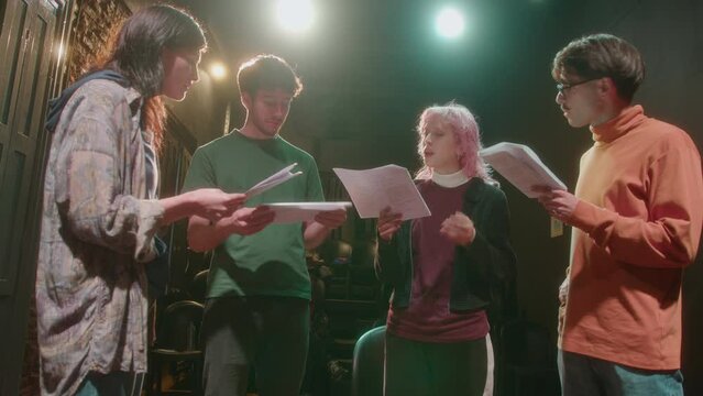 Group of young actors standing on stage with a director and reading scripts from papers during rehearsal or acting class in theater
