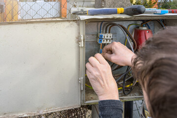 An electrician connects wires in a switchboard outdoors. The hands of an electrician close-up inserts the wire into the attachment point in the electrical panel.