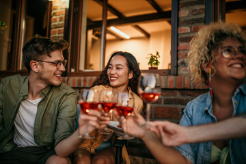 Group of smiling friends sitting and cheering with wine glasses together during a evening on the house porch.